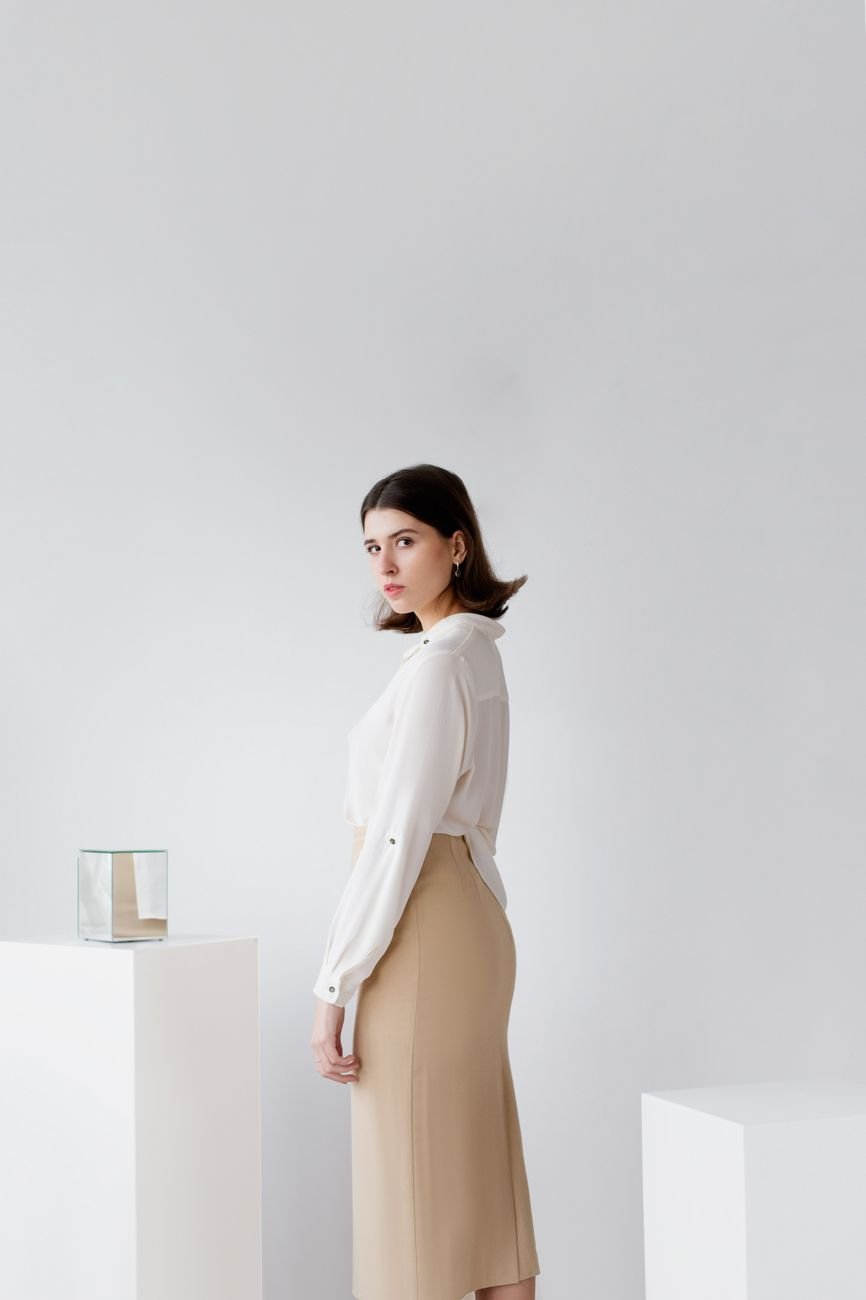 Woman posing indoors in a minimalist setting wearing a white blouse and beige skirt.