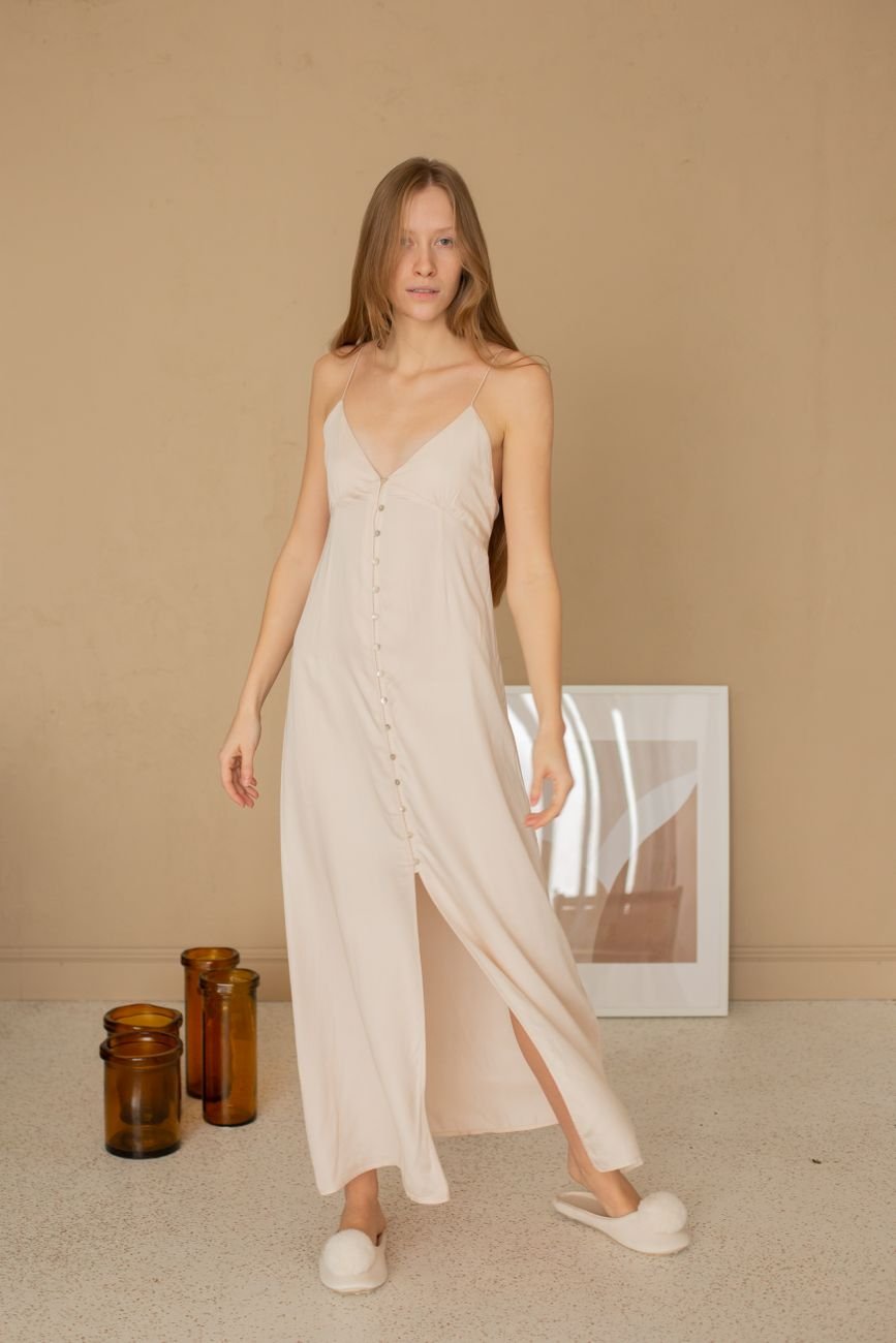 Woman in a beige maxi dress elegantly posing indoors with minimalistic decor.